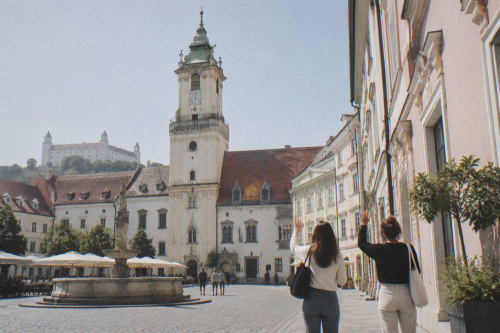 Two friends waving in greeting beside a pastel building facade in Bratislava