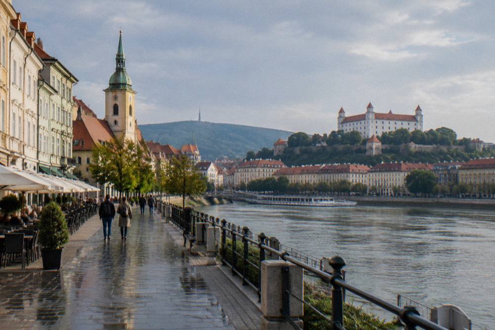 A riverside promenade in Bratislava along the Danube with the castle hill rising behind the city rooftops