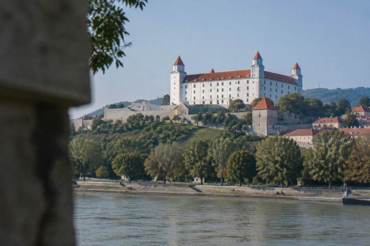 Bratislava Castle seen from the riverbank below, with trees along the Danube promenade in bright daylight.