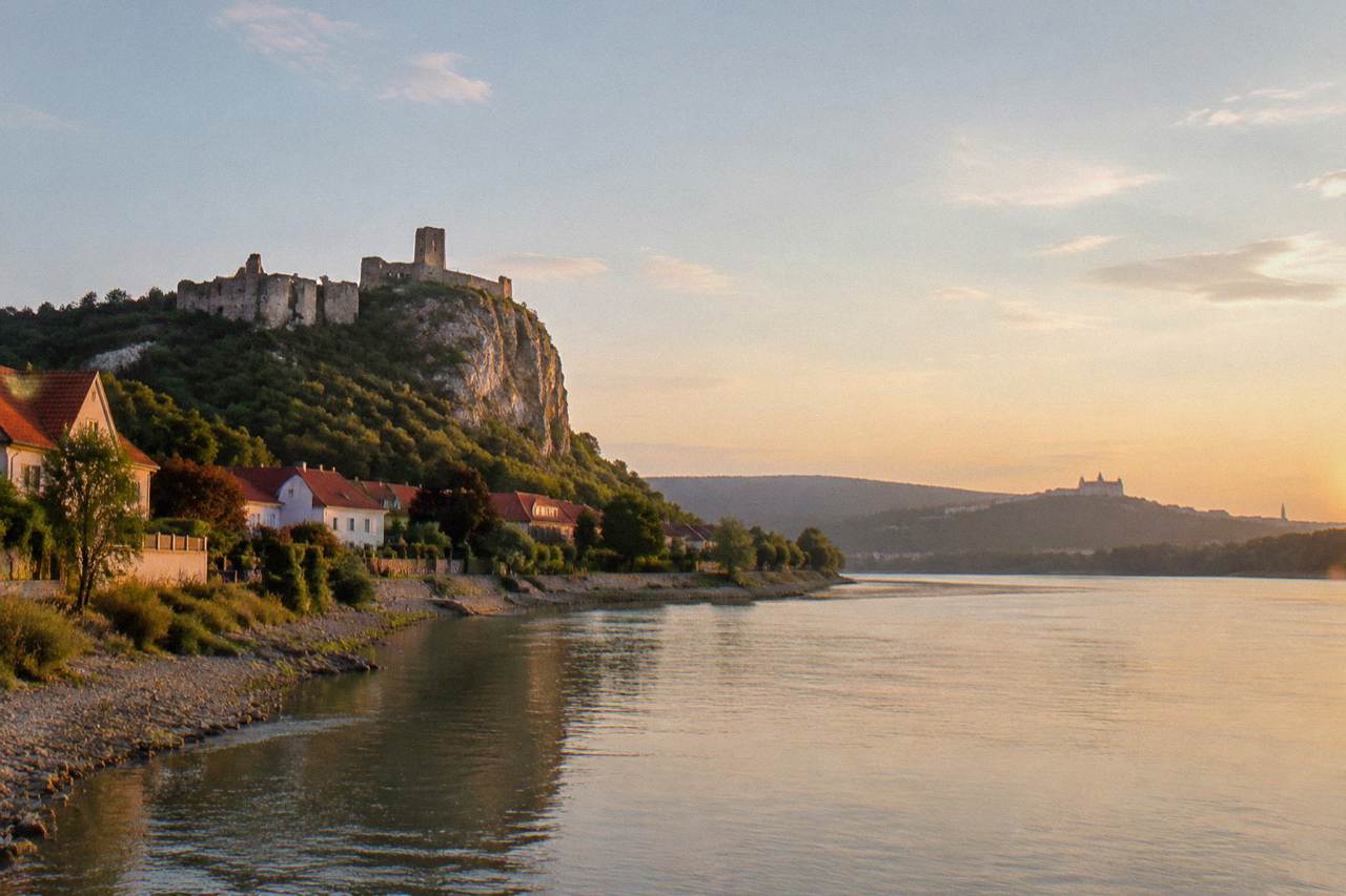Stone walls of a hilltop castle ruin above a wide river with forested slopes behind it