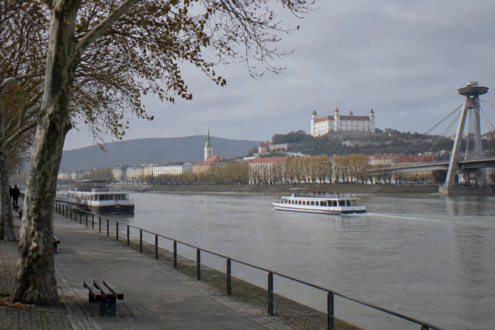 A passenger boat cruising down a wide river past a city embankment with trees