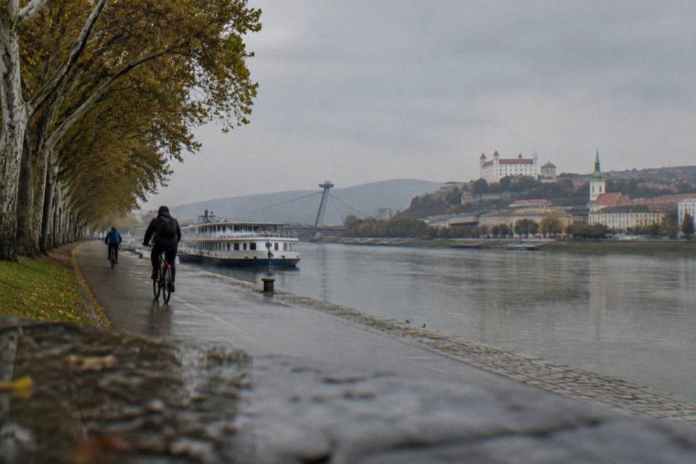 Cyclists riding a flat riverside path with the river on one side and trees on the other