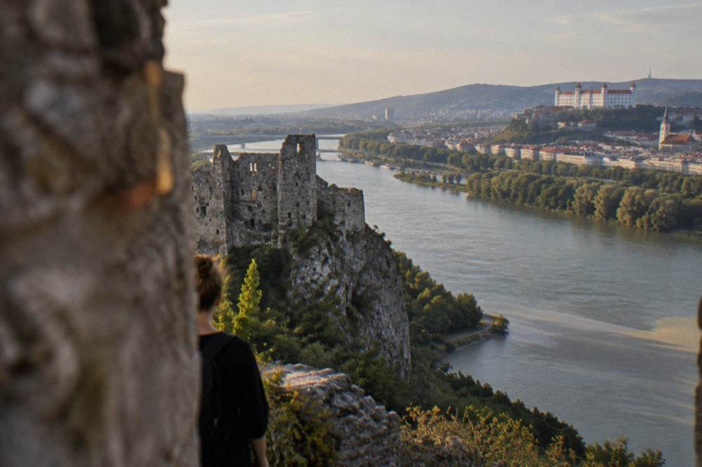 Two rivers joining below a wooded cliff with ruined castle walls on top