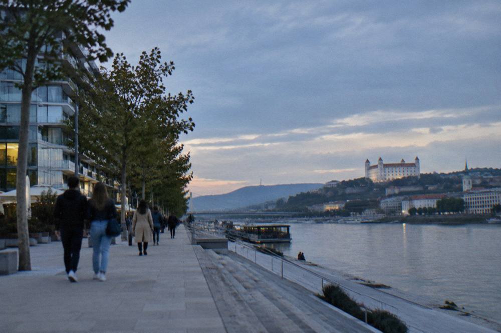 People walking along a Danube riverside walkway with modern buildings behind them in Bratislava