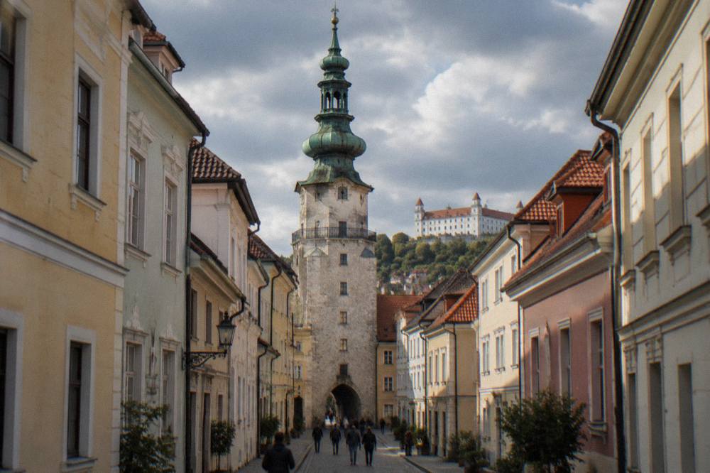 Looking up along a narrow cobbled street lined with coloured townhouses toward a stone gate tower