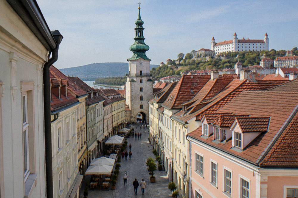 A pedestrian street with cafe tables and pastel facades leading to a stone gate tower