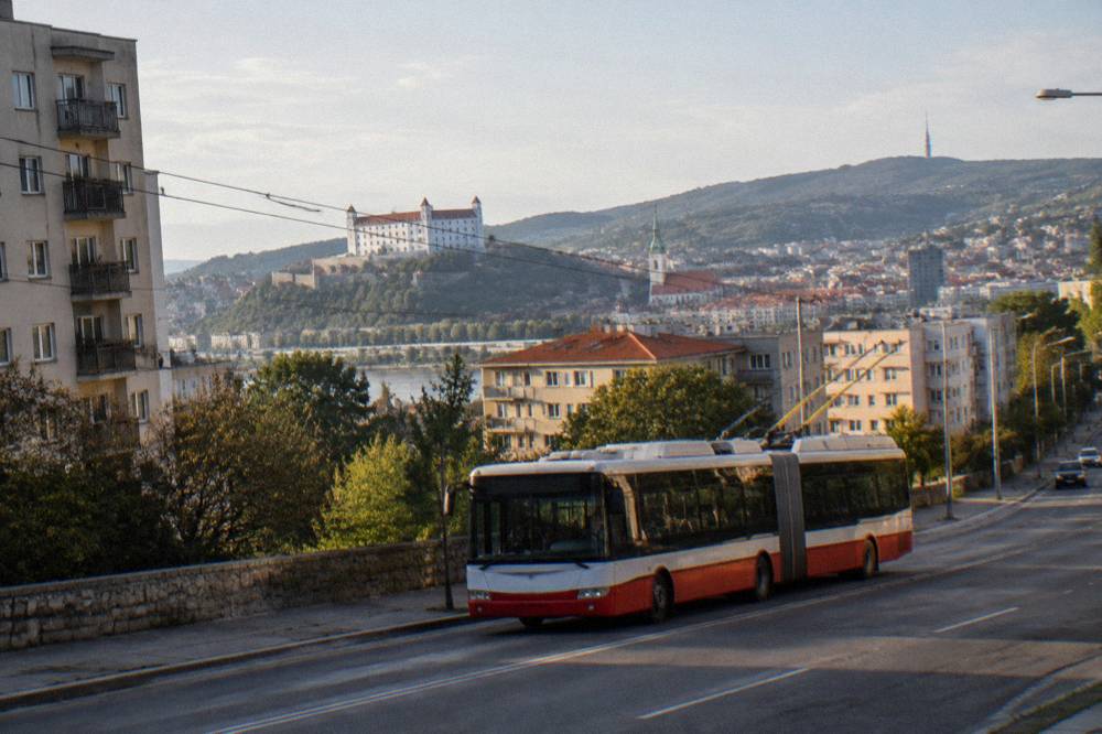 A city trolleybus moving along a street with apartment blocks and green hills in the background