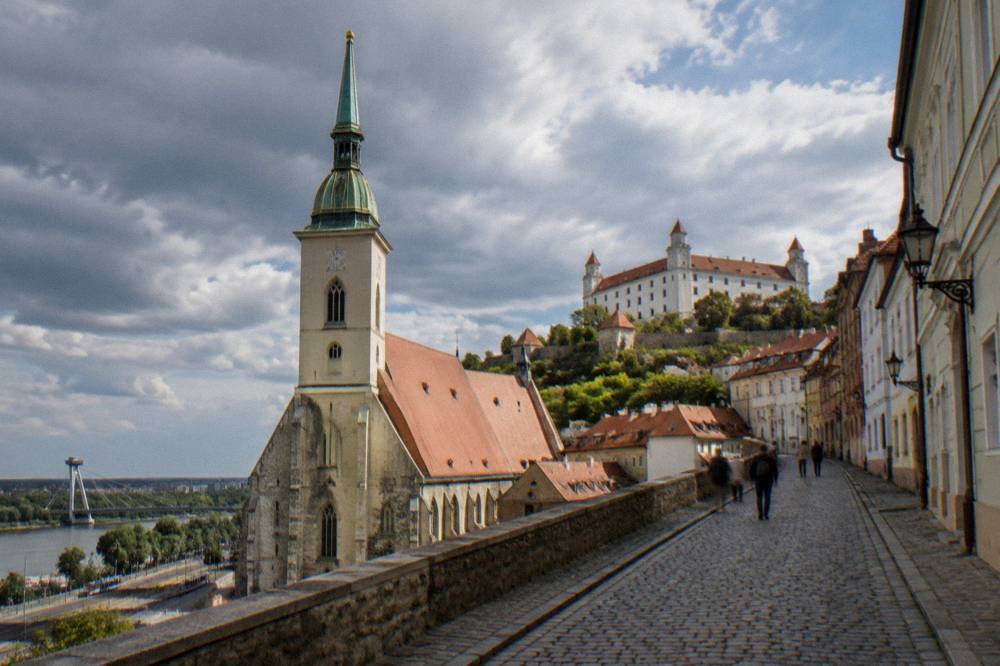 Cobbled street sloping upward from a church toward castle walls on a hill above