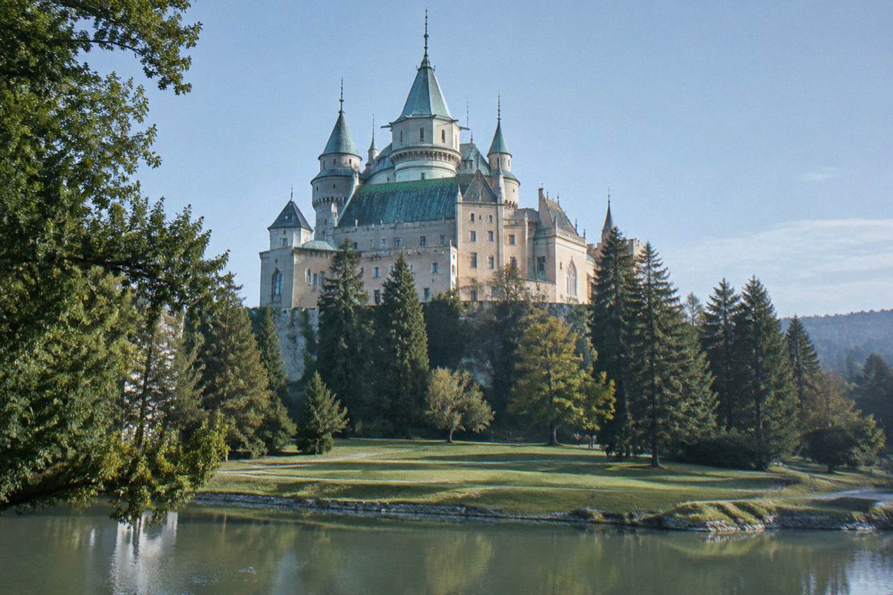 A fairytale-style castle with towers and spires above a park, seen across an open lawn