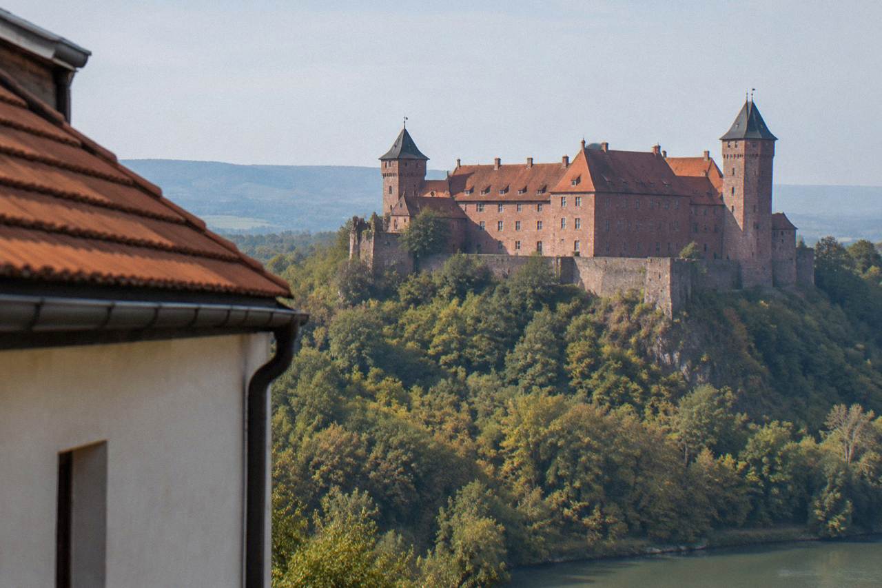 A large red stone castle with corner towers standing on a forested hill above Small Carpathian countryside