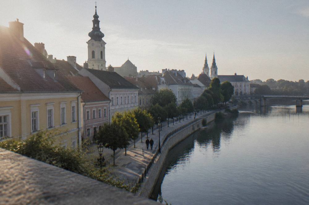 A riverside promenade with church spires behind the rooftops in Győr
