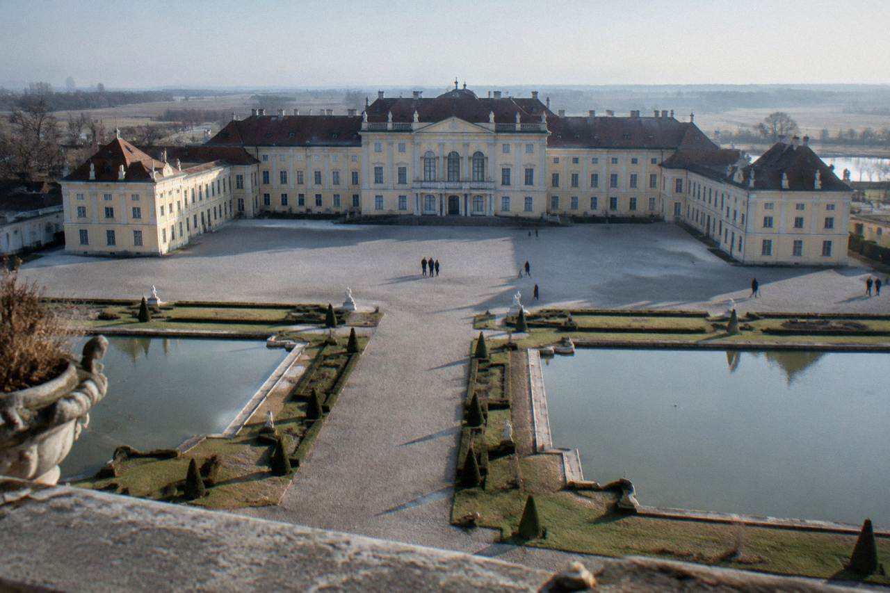 A grand palace facade with symmetrical wings and a wide gravel forecourt