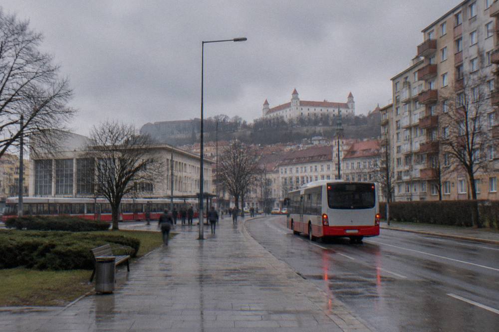 A city bus pulling away from a stop beside a road with apartment blocks behind it