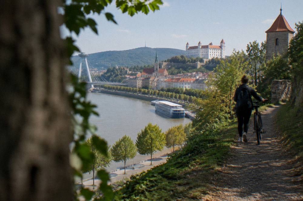 A rider walking a bicycle up a wooded path toward a hilltop tower
