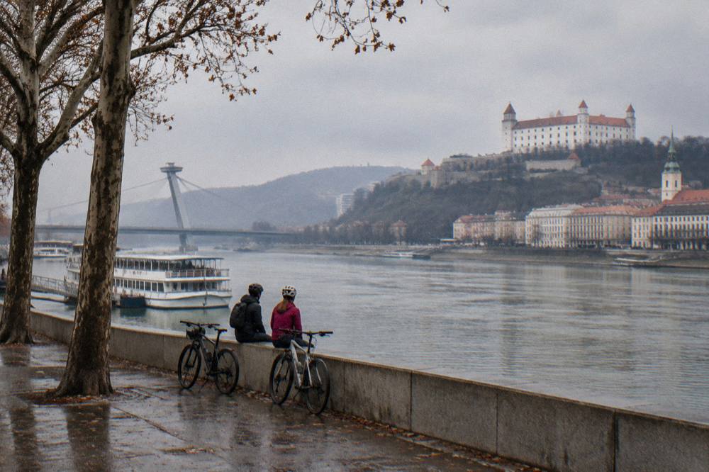Two cyclists resting by the river with a castle on a hill in the distance