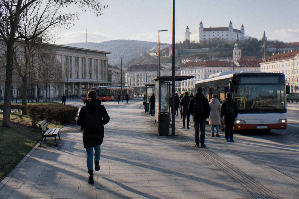 A city bus pulling up to a stop outside a station with travellers waiting