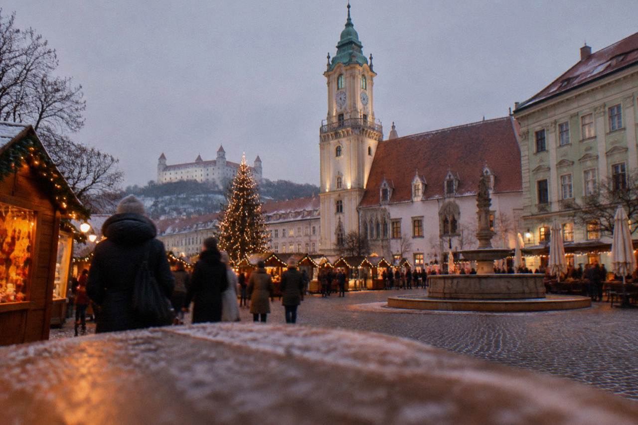 A Christmas market square in Bratislava at dusk with wooden stalls and a lit tree