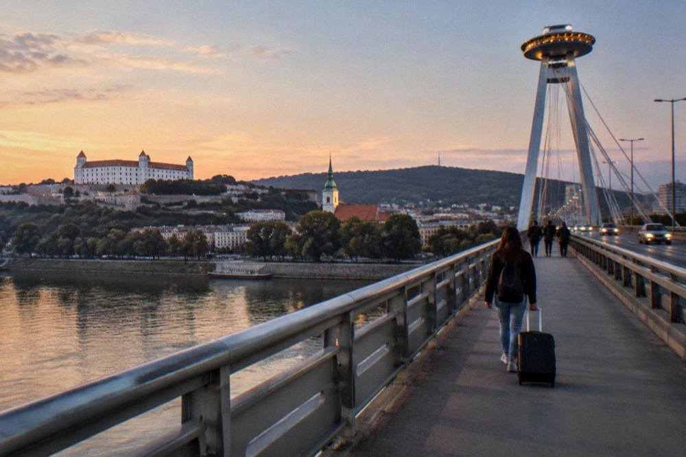 Person pulling a suitcase across a bridge over the Danube at dusk with the illuminated UFO tower in the background