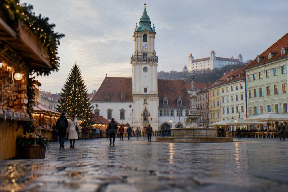 People strolling past market stalls and a decorated tree on a Bratislava square in early evening