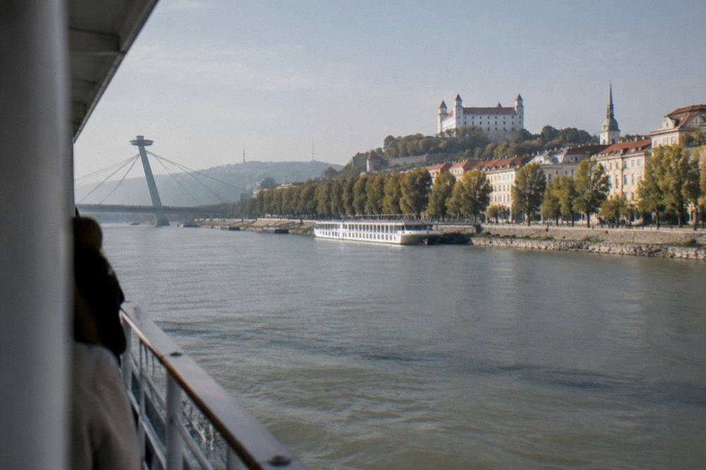 A boat deck view of a bridge pylon and the riverfront promenade along the Danube in Bratislava