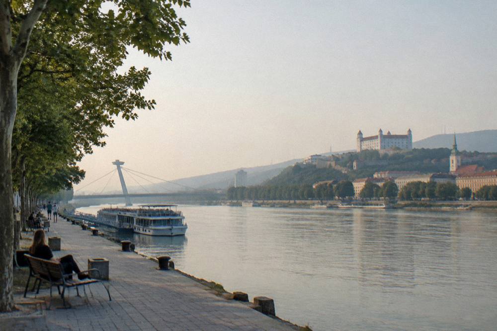 A riverside promenade with trees, benches and a bridge over the Danube at dusk