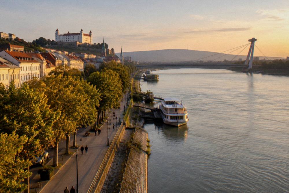 A view along the Danube riverfront toward a bridge with a tower and observation deck