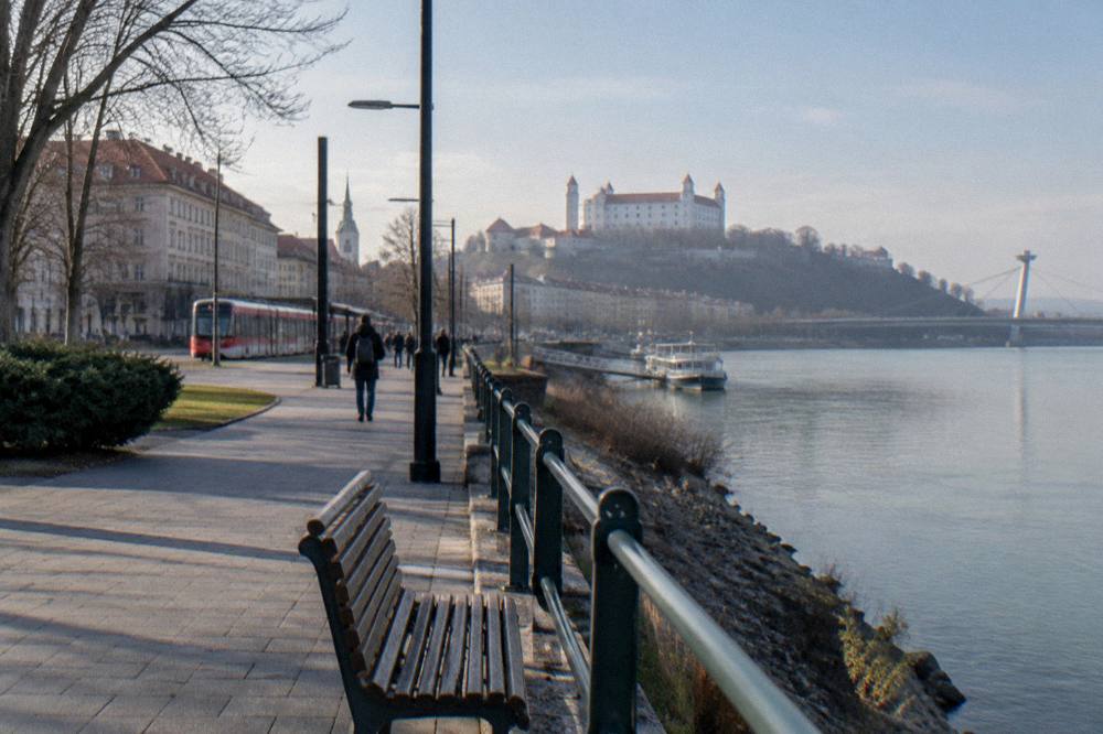 A hilltop castle with white walls and red roofs seen from a riverside path