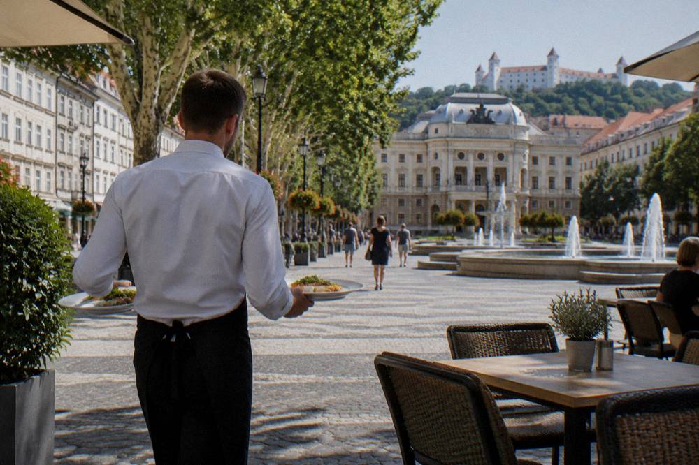 A waiter carrying two plates of food to an outdoor table on a cobbled square