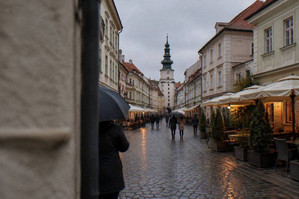 Bratislava Old Town square at dusk with umbrella-carrying pedestrians and lit café windows