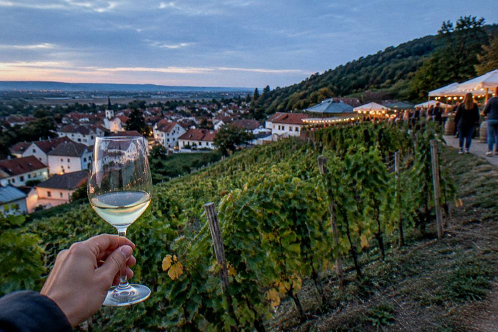 A glass of young white wine raised against an evening market glow